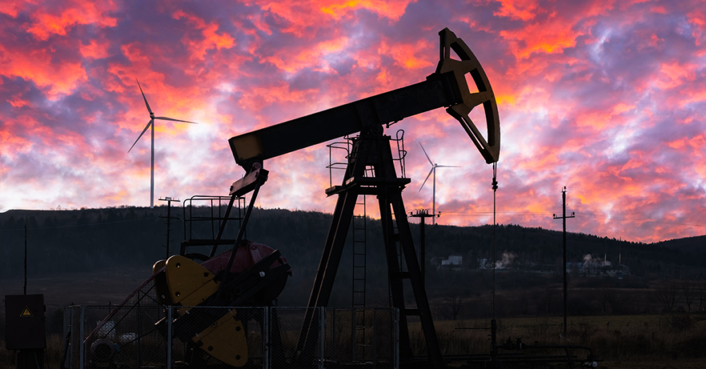 Oil pumpjack silhouetted against a vibrant sunset sky with wind turbines in the background, symbolizing the evolving energy landscape driven by edge computing.