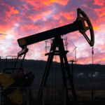 Oil pumpjack silhouetted against a vibrant sunset sky with wind turbines in the background, symbolizing the evolving energy landscape driven by edge computing.