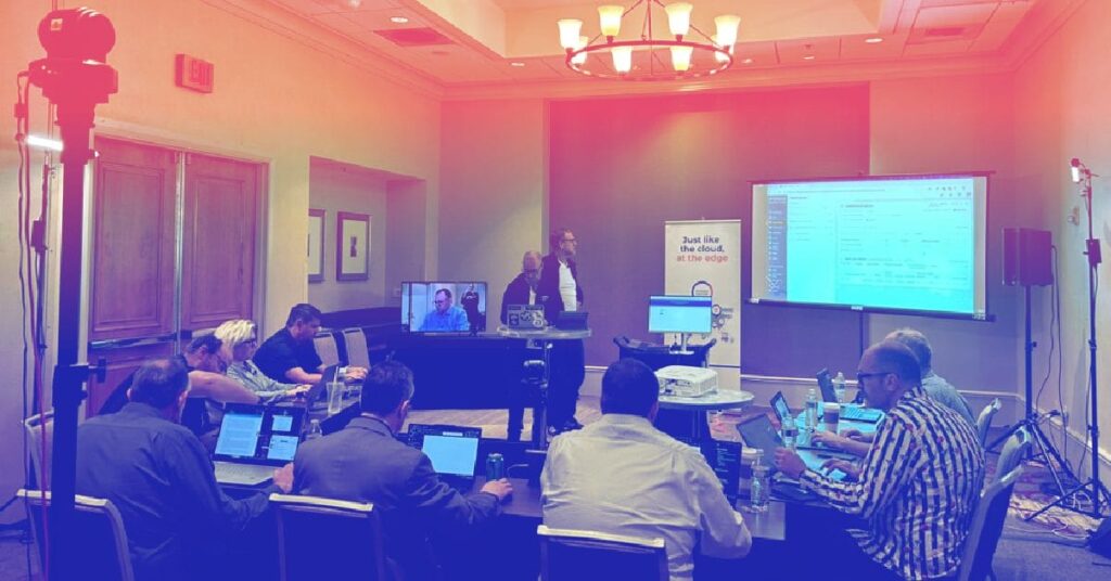 Business conference with attendees using laptops, a presenter speaking, and a screen displaying code.