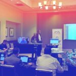 Business conference with attendees using laptops, a presenter speaking, and a screen displaying code.